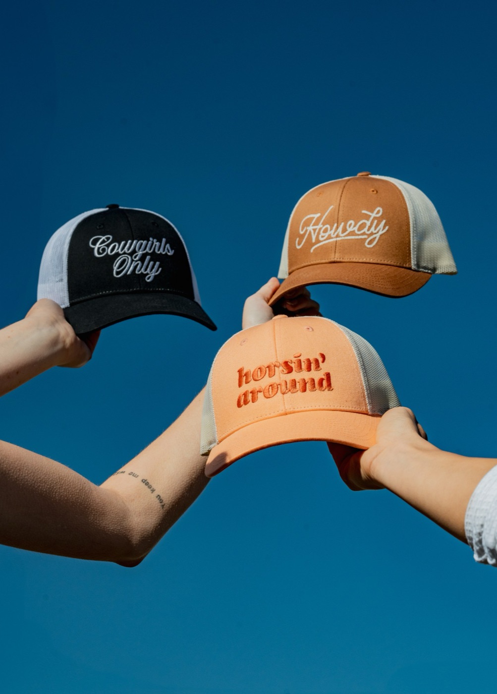 Cowgirls Only, Howdy, and Horsin' Around caps held up against a blue sky