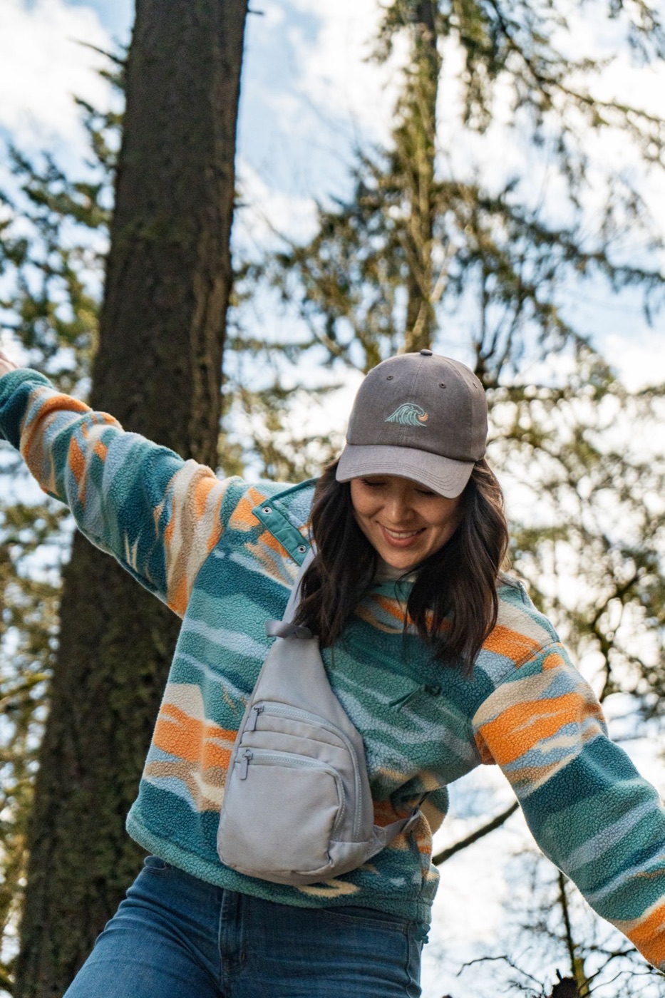 Model in a patterned fleece and wave cap, arms out, under tall trees