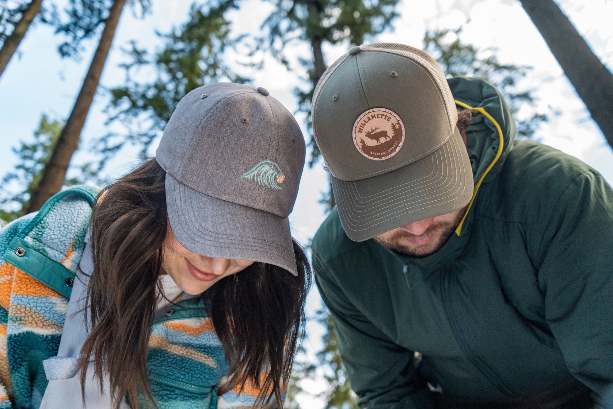Two models in a wave cap and Willamette trucker looking down, shot from below among tall trees