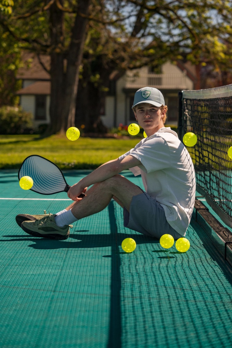 Player in a racquet club cap sitting by the net with pickleballs suspended in the air