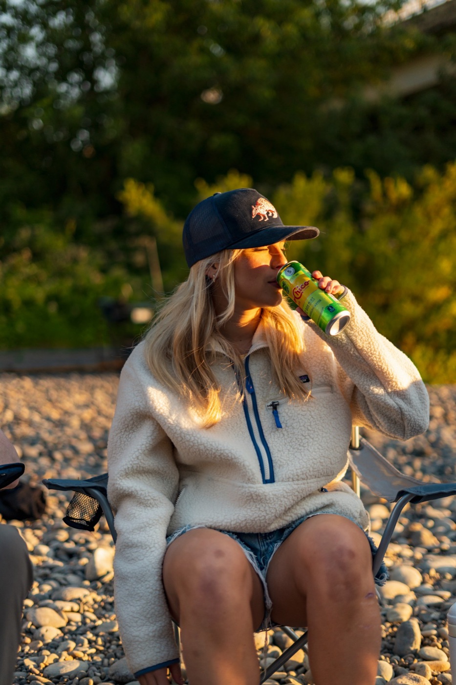 Woman in a navy trucker and fleece sipping a drink in golden light