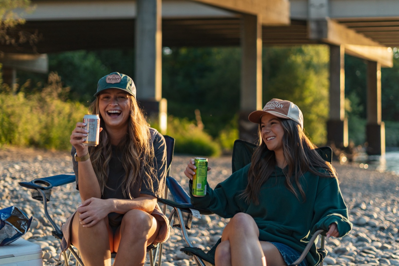Two friends laughing in camp chairs under a bridge