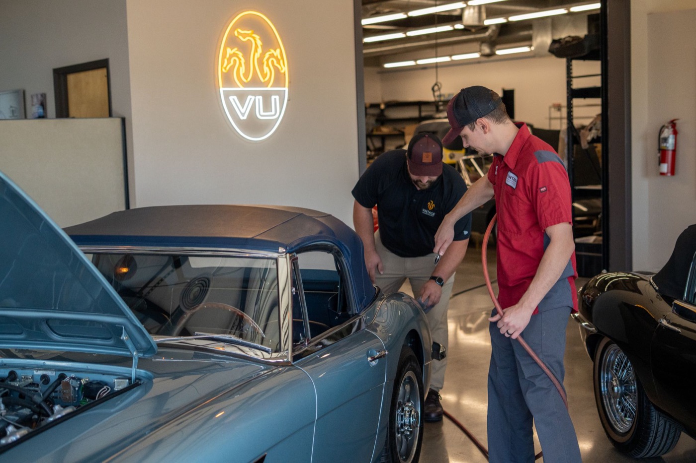Two mechanics inspecting a classic convertible under a neon VU sign